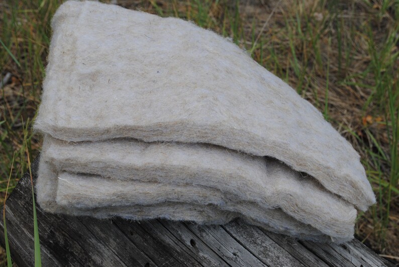 Pode incluir: Uma pilha de material fibroso bege claro, possivelmente isolamento ou estofamento, repousa sobre uma superf&iacute;cie de madeira envelhecida. O material parece macio e em camadas, com uma textura natural e org&acirc;nica. O fundo apresenta grama verde e folhagem.