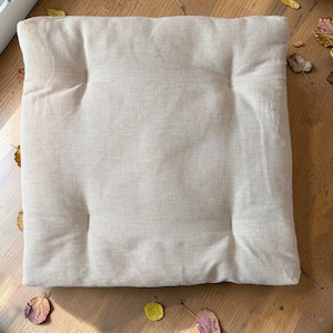 Puede incluir: Un cojín cuadrado beige claro con un diseño acolchado. El cojín está hecho de una tela texturizada y parece estar relleno de un material suave. El cojín está colocado sobre una superficie de madera con hojas de otoño dispersas.