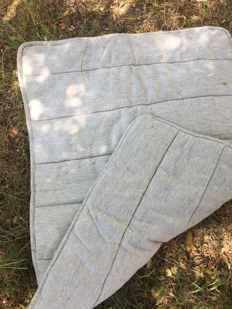 K&ouml;nnte beinhalten: Ein grauer gesteppter Matratzenauflage mit einer strukturierten Oberfl&auml;che. Die Auflage ist in der H&auml;lfte gefaltet und liegt auf einem Bett aus trockenem Gras.