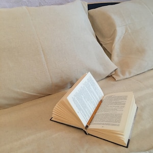 Könnte beinhalten: Ein offenes Buch mit einem Bleistift-Lesezeichen liegt auf einem Bett mit zwei beigen Kissen.