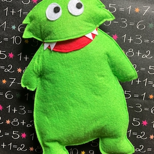 Puede incluir: Un monstruo de fieltro verde con una gran sonrisa de dientes y ojos blancos. El monstruo está sentado sobre un fondo negro con números y estrellas blancas.
