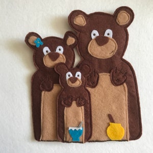 May include: Three brown felt bears of different sizes, the largest bear has a yellow felt pot with a brown felt spoon, the middle bear has a blue felt cup with a white felt straw, and the smallest bear has a blue felt flower on its head.