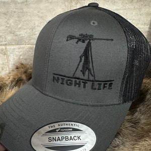 May include: A gray and black mesh baseball cap with a black embroidered design of a rifle and a deer. The cap has a black embroidered text that says "NIGHT LIFE". The cap has a silver label that says "THE AUTHENTIC YUPOONG SNAPBACK YUPOONG SINCE 1974".