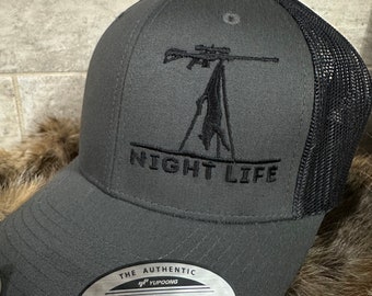 May include: A grey and black mesh baseball cap with a black embroidered design of a rifle and a deer. The cap has a black embroidered text that says "NIGHT LIFE". The cap has a silver label that says "THE AUTHENTIC YUPOONG SNAPBACK YUPOONG SINCE 1974".