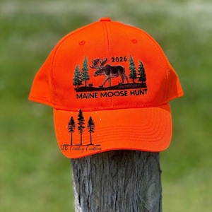 May include: A bright orange baseball cap featuring an embroidered black graphic of a moose standing among pine trees with the text 2026 and MAINE MOOSE HUNT above the brim. The brim displays three black pine trees and the text ShB Country Creations.