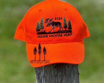 Peut inclure: Une casquette de baseball orange vif avec une broderie noire représentant un élan parmi des pins, accompagnée du texte 2026 et MAINE MOOSE HUNT au-dessus de la visière. La visière présente trois pins noirs et le texte ShB Country Creations.