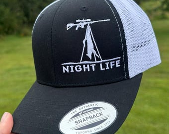 May include: Black and white trucker hat with a white embroidered design of a rifle and a deer on a tripod with the text "NIGHT LIFE". The hat has a black front panel and a white mesh back panel. The hat has a black YUPOONG label on the underside of the brim with the text "THE AUTHENTIC SNAPBACK YUPOONG SINCE 1974".