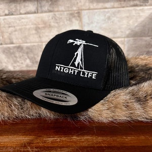 May include: Black and white embroidered trucker hat with a silhouette of a person holding a rifle and the text "NIGHT LIFE". The hat has a black mesh back and a black front with a white "THE AUTHENTIC SNAPBACK YUPOONG SINCE 1974" label.