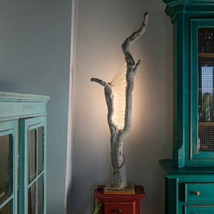May include: A unique floor lamp made from a natural driftwood branch with a string of lights woven through the branches. The lamp is on a red wooden base and is lit, casting a warm glow.