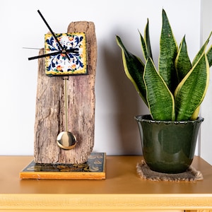 May include: A clock with a blue, yellow and white patterned face is mounted on a piece of driftwood. The clock has a pendulum and is sitting on a table with a potted snake plant.