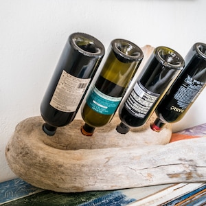 Puede incluir: Un rústico estante de vino de madera hecho de madera a la deriva sostiene cuatro botellas de vino. El estante es de color marrón claro natural con una textura rugosa y desgastada. Las botellas son una mezcla de vino tinto y blanco.