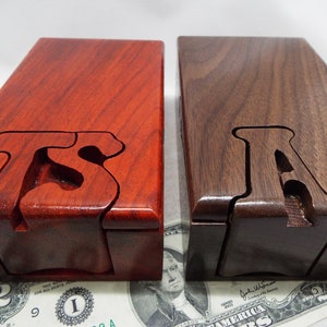 May include: Two wooden puzzle boxes, one red and one brown, with the letter "A" carved into the top of each box. The boxes are sitting on a dollar bill.