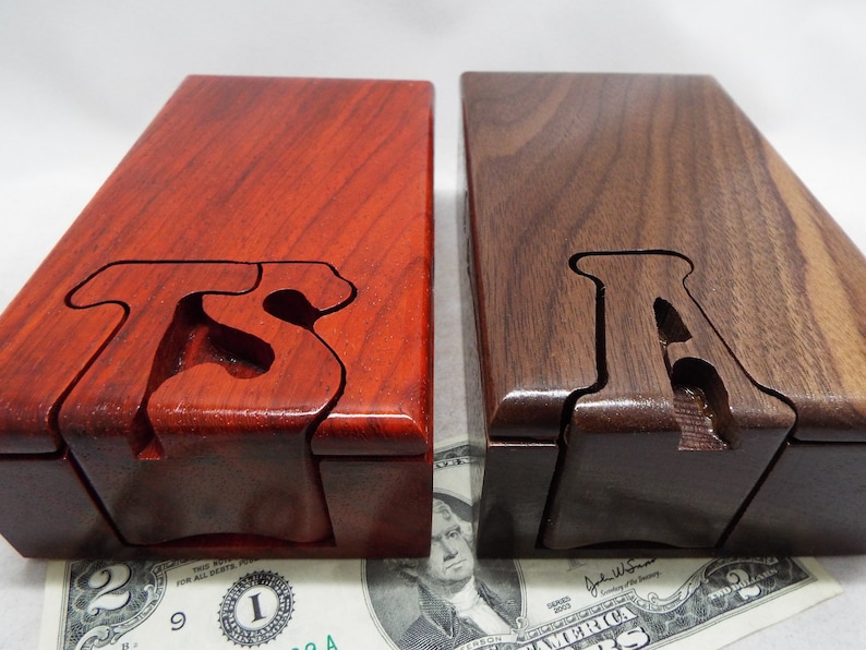 May include: Two wooden puzzle boxes, one red and one brown, with the letter "A" carved into the top of each box. The boxes are sitting on a dollar bill.