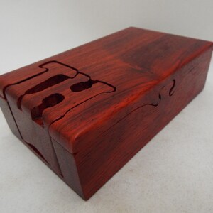 May include: A red wooden box with a unique interlocking puzzle design. The box is made of a dark red wood with a natural grain pattern.
