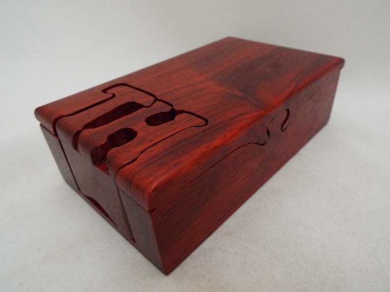 May include: A red wooden box with a unique interlocking puzzle design. The box is made of a dark red wood with a natural grain pattern.