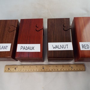 May include: Four wooden boxes with heart-shaped cutouts, each made from a different type of wood: mahogany, padauk, walnut, and red cedar. The boxes are arranged in a row from left to right, with a ruler below them.