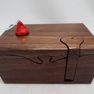 May include: A wooden box with a heart-shaped cutout and a puzzle-like lid. The box is made of dark wood and has a smooth finish. A red chocolate kiss is sitting on top of the box.
