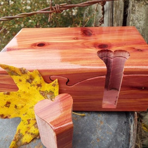 May include: A wooden heart-shaped puzzle box with a cedar wood finish. The box is open and the lid is resting on top of the box. A heart-shaped piece is missing from the lid. A fall leaf is lying next to the box.