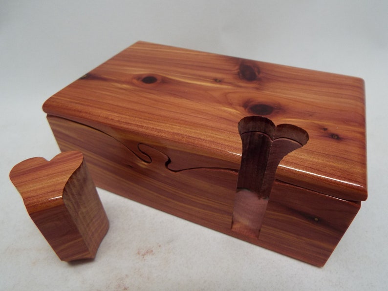 May include: A wooden puzzle box with a heart-shaped lid. The box is made of cedar wood and has a natural reddish-brown color. The lid is hinged and opens to reveal a small compartment inside.