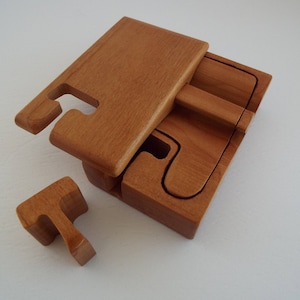 May include: A wooden puzzle box with a lid that lifts off. The box is made of light brown wood and has a natural finish. The box is open and the lid is lying on top of the box. There is a small wooden piece that fits into the box.