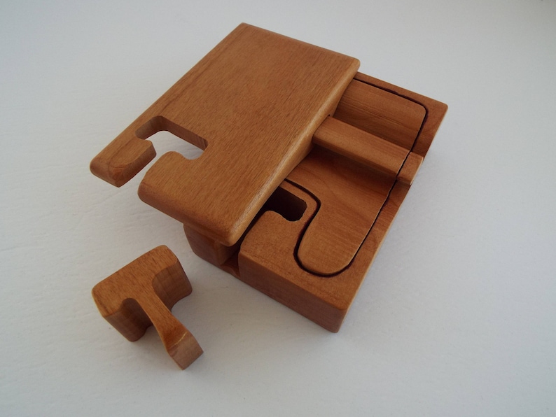 May include: A wooden puzzle box with a lid that lifts off. The box is made of light brown wood and has a natural finish. The box is open and the lid is lying on top of the box. There is a small wooden piece that fits into the box.