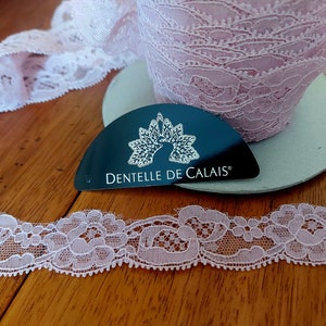Peut inclure: Galon de dentelle rose avec un bord festonné. La dentelle est délicate et présente un motif floral. L'étiquette indique "Dentelle de Calais".