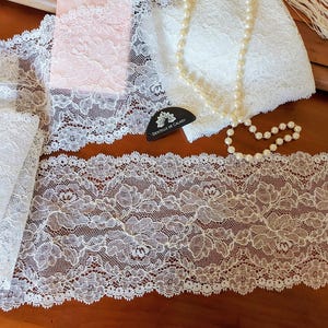 Può includere: Primo piano di vari tessuti di pizzo bianchi e rosa, con una collana di perle e un'etichetta con la scritta "Dentelle de Calais". Il pizzo presenta motivi floreali e trame delicate, ideale per cucito e progetti creativi.