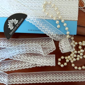 Peut inclure: Plusieurs rouleaux de dentelle blanche et un collier de perles sont présentés sur une surface en bois. La dentelle présente un bord festonné délicat et un motif géométrique répétitif. Une étiquette noire avec "Dentelle de Calais" est visible.