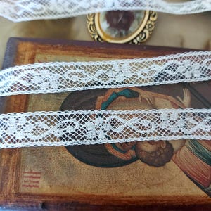 Peut inclure: Une longueur de dentelle blanche ornée d'un motif floral délicat sur un fond en maille hexagonale. La dentelle est drapée sur une icône en bois rectangulaire représentant une figure religieuse, avec un petit cadre ovale doré visible au-dessus.