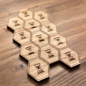 Peut inclure: Un ensemble de 10 pièces de jeu hexagonales en bois avec un symbole de sablier gravé sur chaque pièce. Les pièces sont disposées en forme de nid d'abeille.