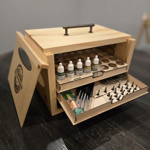 Puede incluir: Una caja de madera con un asa en la parte superior, que contiene pequeños frascos de pintura y un cajón con herramientas y figuras en miniatura. La caja está hecha de madera clara y tiene un acabado natural.