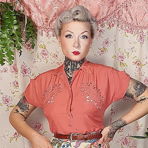 May include: A short-sleeved coral button-down shirt with embroidered detailing on the chest, paired with a cream-colored skirt featuring a vibrant multi-colored floral pattern, a thin brown belt, and light-colored shoes with a cross-strap design.