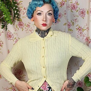 May include: A woman wearing a light yellow cardigan with a scalloped edge, paired with a floral print skirt featuring pink, blue, and yellow flowers on a black background. The outfit is completed with tan shoes.