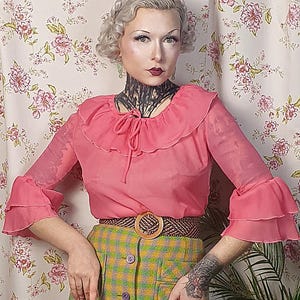 Könnte beinhalten: Eine Frau trägt eine rosa Bluse mit Rüschenkragen und einen grün-gelben karierten Rock mit einer Reihe von Knöpfen vorne. Sie trägt einen braunen Gürtel mit einer großen goldenen Schnalle.