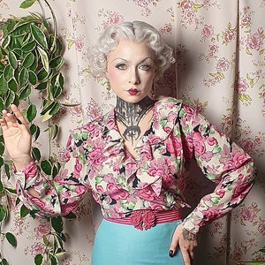 Può includere: Una donna indossa una blusa vintage a stampa floreale con colletto a volant e una gonna turchese. È in piedi davanti a uno sfondo con carta da parati floreale e una pianta in vaso.