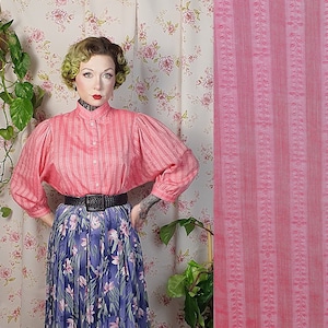 May include: A pink and white striped blouse with puffed sleeves and a high collar. The blouse is worn with a black belt and a floral skirt.
