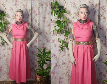 May include: A pink sleeveless dress with a gold belt. The dress has a high neckline and a full skirt. The model is wearing white gloves.