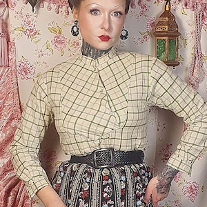 Könnte beinhalten: Ein Outfit im Vintage-Stil mit einer karierten Bluse, einem floral bedruckten Rock und einem schwarzen Gürtel. Der Rock hat einen schwarzen Hintergrund mit vertikalen Streifen und roten, weißen und blauen Blumenmustern. Das Outfit wird durch gemusterte Absätze ergänzt.