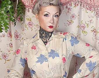 May include: A long-sleeved button-down shirt with a cream base and a blue and pink floral pattern, paired with a tan knee-length skirt featuring two front pockets and a center pleat, finished with a thin white belt and white peep-toe heels.