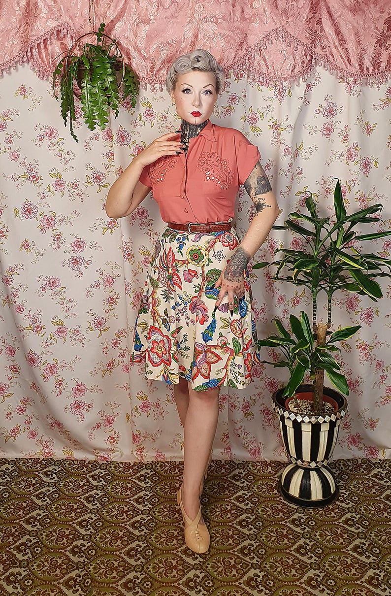 May include: A short-sleeved coral button-down shirt with decorative embroidery on the chest, paired with a cream-colored A-line skirt featuring a vibrant multi-colored floral pattern, a thin brown leather belt, and light beige closed-toe shoes.