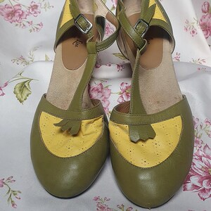 Op de afbeelding: Een paar olijfgroene en gele T-band schoenen. De schoenen hebben een ronde neus en een kleine hak. De T-band en de enkelband hebben een gespsluiting. De schoenen worden gepresenteerd op een stof met bloemenpatroon.