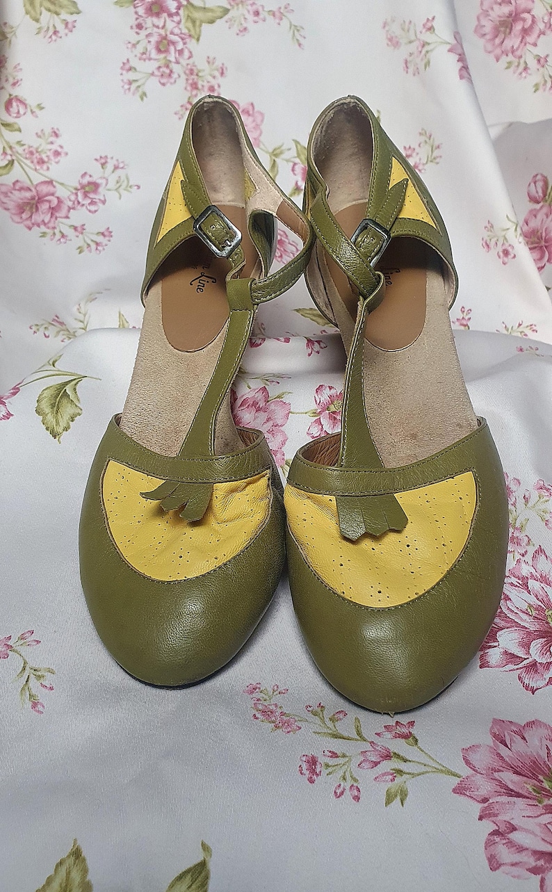 Op de afbeelding: Een paar olijfgroene en gele T-band schoenen. De schoenen hebben een ronde neus en een kleine hak. De T-band en de enkelband hebben een gespsluiting. De schoenen worden gepresenteerd op een stof met bloemenpatroon.