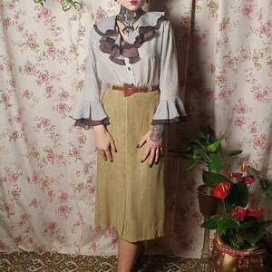 May include: A woman wearing a white blouse with black polka dots and brown ruffles, a brown suede skirt, and brown leather belt. She is standing in front of a floral patterned wall with a potted plant to the right.