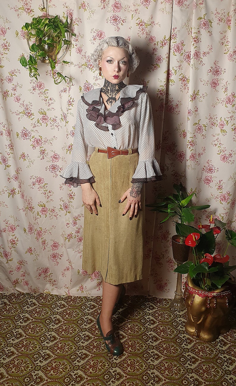 May include: A woman wearing a white blouse with black polka dots and brown ruffles, a brown suede skirt, and brown leather belt. She is standing in front of a floral patterned wall with a potted plant to the right.