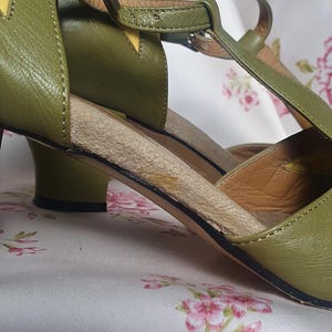 Op de afbeelding: Olijfgroene leren sandalen met een T-strap design. De sandalen hebben een kleine hak en een gespsluiting. Gele accenten zijn zichtbaar op de bovenkant. De schoenen staan tegen een bloemenachtergrond.