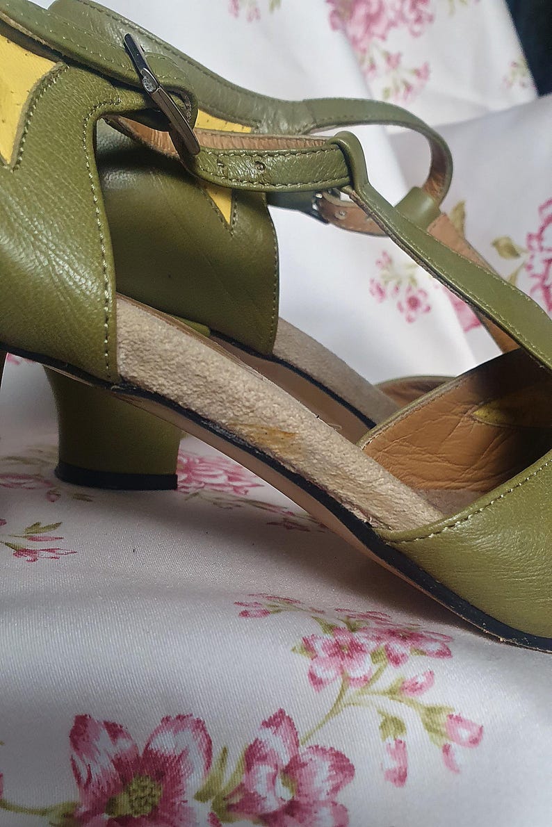 Op de afbeelding: Olijfgroene leren sandalen met een T-strap design. De sandalen hebben een kleine hak en een gespsluiting. Gele accenten zijn zichtbaar op de bovenkant. De schoenen staan tegen een bloemenachtergrond.