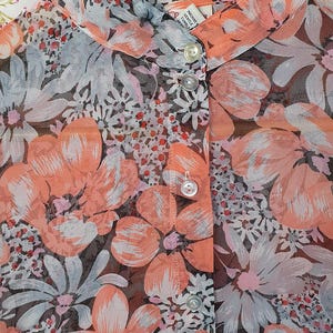 以下が含まれることがあります： 花柄の透き通るピーチ色のブラウス。ブラウスはフロントにボタン留め、襟付き。生地は軽量で繊細な感触。花はピンク、白、グレーの色合いです。