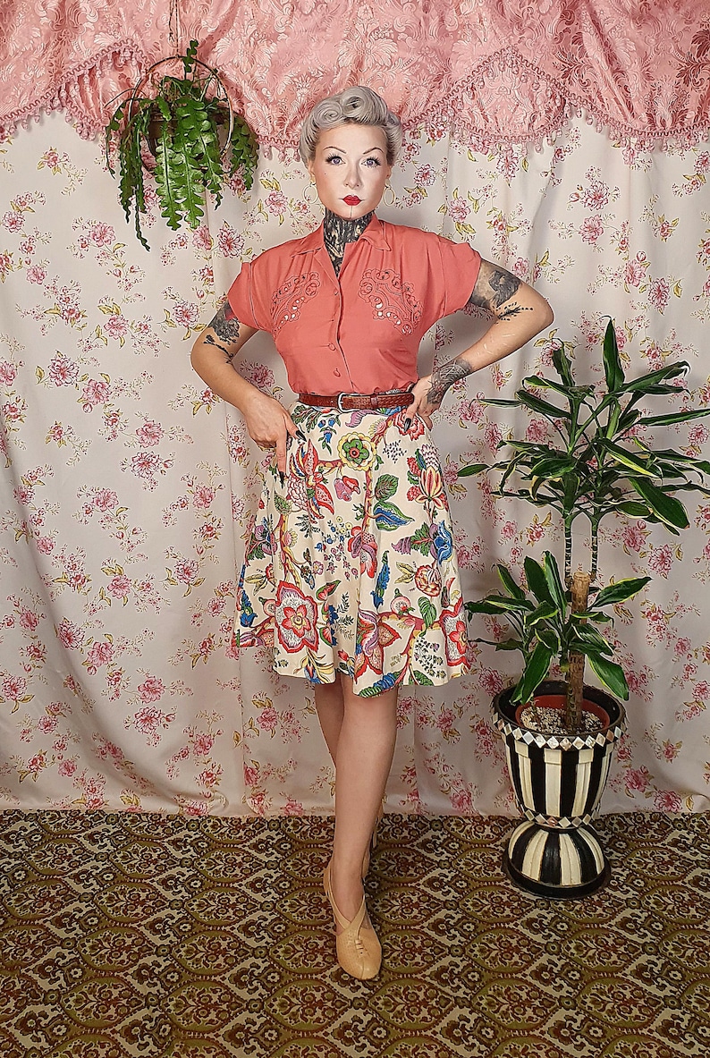 May include: A short-sleeved coral button-down shirt with embroidered detailing on the chest, paired with a cream-colored skirt featuring a vibrant multi-colored floral pattern, a thin brown belt, and light-colored shoes with a cross-strap design.