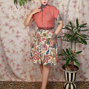 May include: A short-sleeved coral button-down shirt with decorative embroidery on the chest, paired with a cream-colored A-line skirt featuring a vibrant multi-colored floral pattern, a thin brown leather belt, and light beige closed-toe shoes.