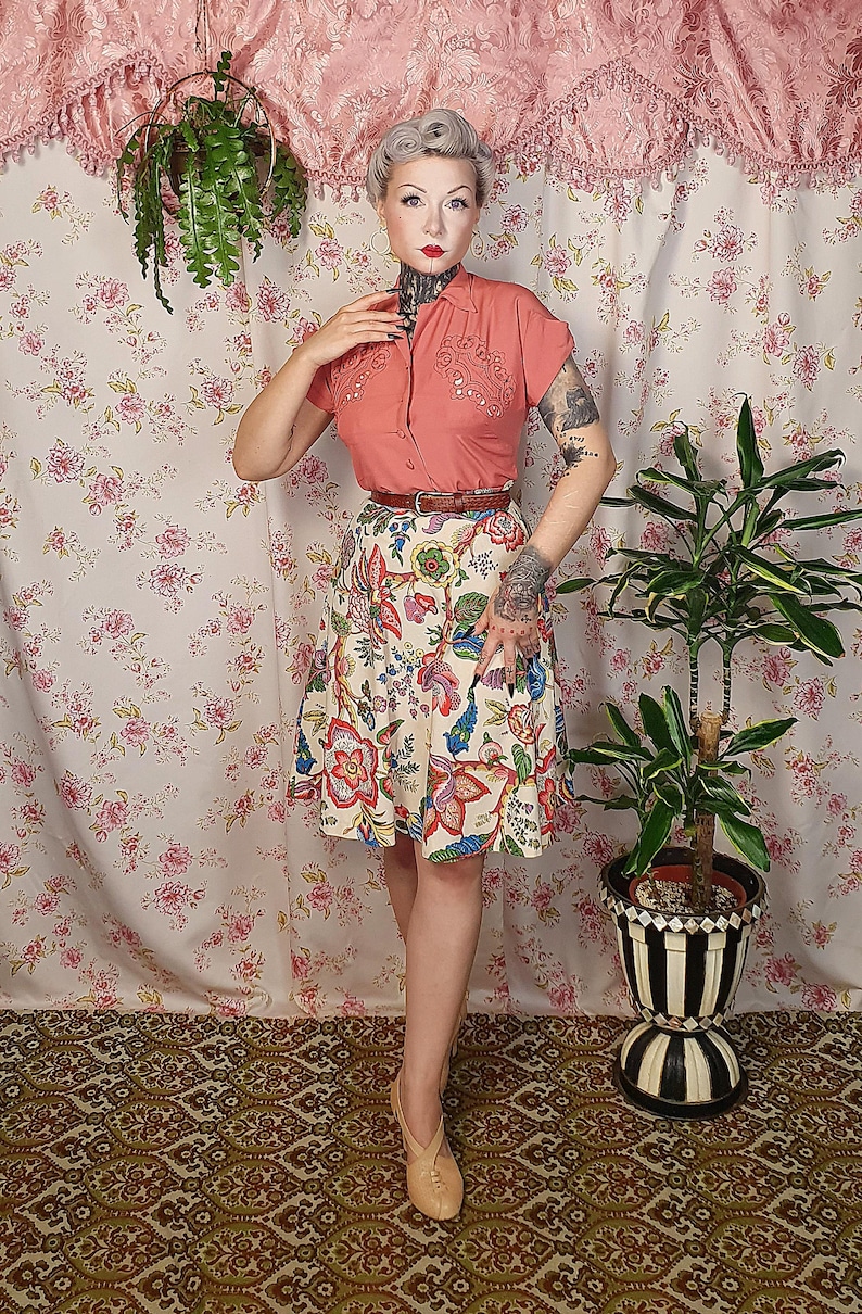 May include: A short-sleeved coral button-down shirt with decorative embroidery on the chest, paired with a cream-colored A-line skirt featuring a vibrant multi-colored floral pattern, a thin brown leather belt, and light beige closed-toe shoes.